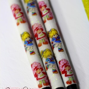 May include: White pens with glitter and Strawberry Shortcake character designs. The pens feature black ink tips and are decorated with images of the classic cartoon character in various outfits. The pens are a nostalgic item.