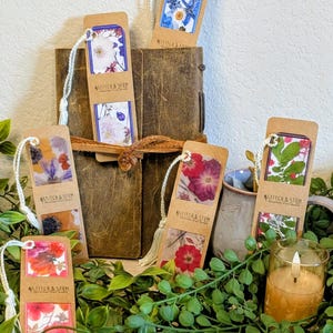 May include: Five rectangular bookmarks made of brown cardstock, each featuring pressed flower designs and a white cord tassel. The text "STITCH & STEM" is printed on each bookmark. The bookmarks are arranged around a brown leather-bound journal.