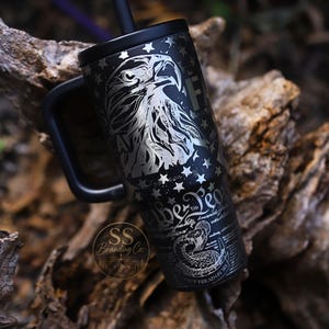 May include: A black stainless steel tumbler with a handle and straw, featuring a laser-engraved design of a bald eagle, stars, and a coiled rattlesnake with the text "We The People" and "Don't Tread On Me" along with a circular logo.