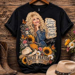 May include: A black t-shirt featuring a graphic of Dolly Parton with blonde hair, a guitar, sunflowers, butterflies, and a cowboy hat. The text reads: If you see someone without a smile... give them one of yours. Dolly Parton 1946-2026.