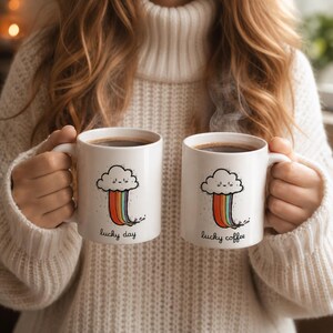 Puede incluir: Dos tazas de cerámica blanca llenas de café, cada una con un diseño de nube de dibujos animados y arcoíris, con las palabras "lucky day" y "lucky coffee". Las tazas son sostenidas por una persona que lleva un suéter blanco.