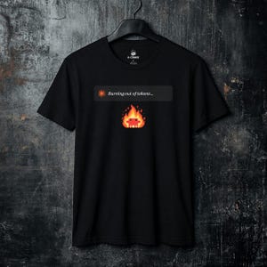 May include: A black short-sleeved t-shirt featuring a graphic print on the chest. The design includes a grey rectangular box with the text "Burning out of tokens..." next to a sun icon, positioned above a pixelated red character engulfed in flames.