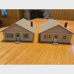 May include: Two miniature model houses with tan roofs, light grey siding, and red brick chimneys. Each house has two windows and a door. The houses are set on a wooden surface.