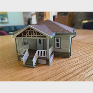 Könnte beinhalten: Ein Miniaturmodellhaus mit hellgrauer horizontaler Fassadenverkleidung, einem braunen Schindeldach und einer Veranda mit weißen Geländern und Treppen. Das Gebäude hat eine weiße Eingangstür mit ovalem Fenster und weiß gerahmte rechteckige Fenster.