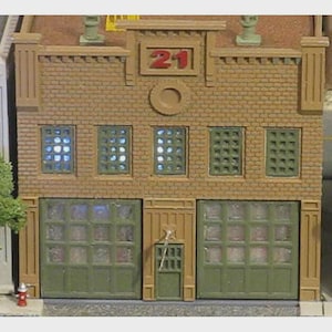 May include: A miniature model of a brick building with the number 21 above the windows. The building has green garage doors and windows, with a small door on the right. The model is detailed with a fire hydrant.
