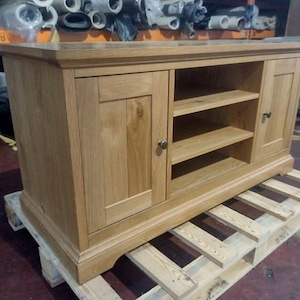 Puede incluir: Un mueble de madera marrón claro con dos puertas y estanterías abiertas. El mueble tiene forma rectangular y está sobre un palé de madera. El mueble tiene un acabado de veta de madera natural y herrajes de color latón.