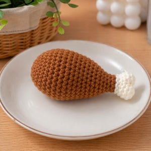 May include: A crocheted brown chicken leg on a white plate. The leg has a white tip and is made of textured yarn. The plate has a gold rim and is set on a wooden surface.