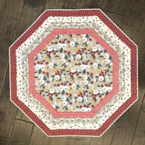 May include: An octagonal quilted table mat featuring a central panel with white and rust-colored floral patterns on a light gray background, surrounded by concentric borders in solid pink, cream with botanical prints, and a dark red polka dot edge.