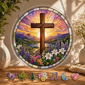 May include: Round stained glass window art featuring a wooden cross set against a sunset over a landscape with mountains, a lake, and flowers. The window is surrounded by a decorative border and has a chain for hanging. Decorative pins are displayed below.