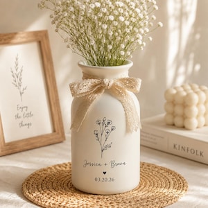 May include: A cream-coloured ceramic vase with a floral design and a lace bow, filled with white flowers. The vase is personalised with the names "Jessica + Brown" and the date "03.20.26". A framed print with the text "Enjoy the little things" is visible.