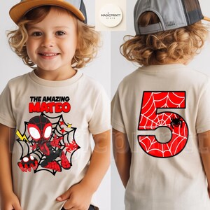 May include: A tan t-shirt featuring a cartoon Spider-Man design with the text "THE AMAZING MATEO" in red and black. The back of the shirt displays a large red number 5 with a spiderweb design and a black spider. The shirt is for a child.