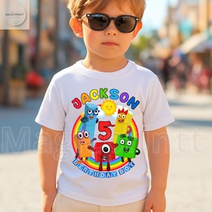 May include: White t-shirt with a colorful graphic design for a child's birthday. The design includes the name "Jackson", the number "5", and the words "Birthday Boy" with cartoon characters and a rainbow.