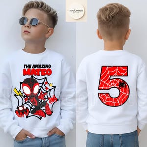 May include: White sweatshirt featuring a graphic of a superhero with the text "THE AMAZING MATEO" on the front. The back of the sweatshirt displays a large red number "5" with a spiderweb design and a small spider.