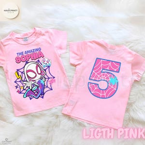 May include: Two light pink t-shirts. One features a cartoon character and the text "THE AMAZING SOPHIA". The other shirt has a large number "5" with a spiderweb design. Both shirts are short-sleeved.