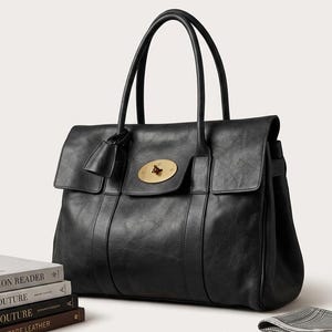 May include: A black leather tote bag with a flap closure and a gold-tone clasp. The bag has two top handles and a small leather tag. The bag is sitting next to a stack of books.