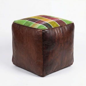 Puede incluir: Un puf cuadrado con base de cuero marrón y parte superior a cuadros en tonos verdes, amarillos, rojos y morados. El puf tiene forma de cubo y parece estar bien construido.