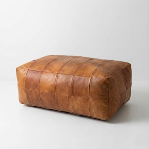 May include: A rectangular, brown leather ottoman with a patchwork design. The ottoman is a warm, medium brown color and appears to be made of individual leather squares stitched together. It is a low, wide piece of furniture.