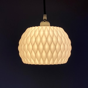 Op de afbeelding: Een hanglamp met een witte, getextureerde lampenkap. De kap heeft een herhalend patroon en hangt aan een zwarte kabel. Het licht is aan en geeft een warme gloed.