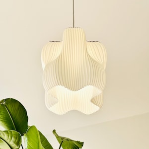Può includere: Una moderna lampada a sospensione bianco sporco con un design unico e stratificato. La luce ha una trama a coste e una forma distintiva, sospesa a un cavo sottile. L'apparecchio è illuminato, proiettando una luce soffusa.