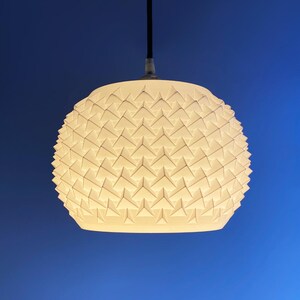 May include: A white, geometric pendant light fixture with a textured surface. The light hangs from a black cord against a blue background. The shade has a pattern of repeating triangular shapes.
