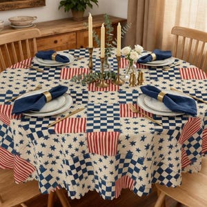 May include: A round tablecloth with a patchwork design featuring blue stars, red stripes, and blue and white checkered squares. The table is set with blue napkins, gold napkin rings, and gold silverware. Candles and flowers are in the center.