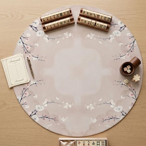 May include: A round, light beige mahjong mat featuring a delicate cherry blossom branch pattern. The set includes two wooden tile racks, a scorecard pad with a pen, a cylindrical dice cup with two dice, a stack of chips, and a box of mahjong tiles.