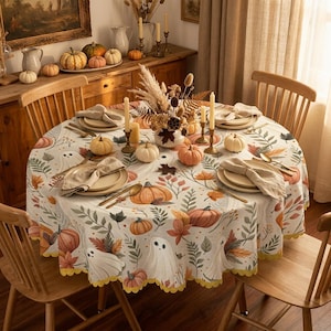 May include: A round tablecloth featuring a pattern of white ghosts, orange pumpkins, and autumn leaves in shades of brown, green, and yellow. The tablecloth has a scalloped yellow edge and is set with plates, napkins, and small white pumpkins.