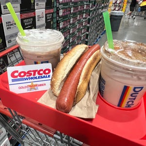 May include: A red shopping cart holds two iced coffee drinks with green straws, a hot dog in a bun, and a Costco Gold Star Member card. Coffee boxes are stacked in the background.