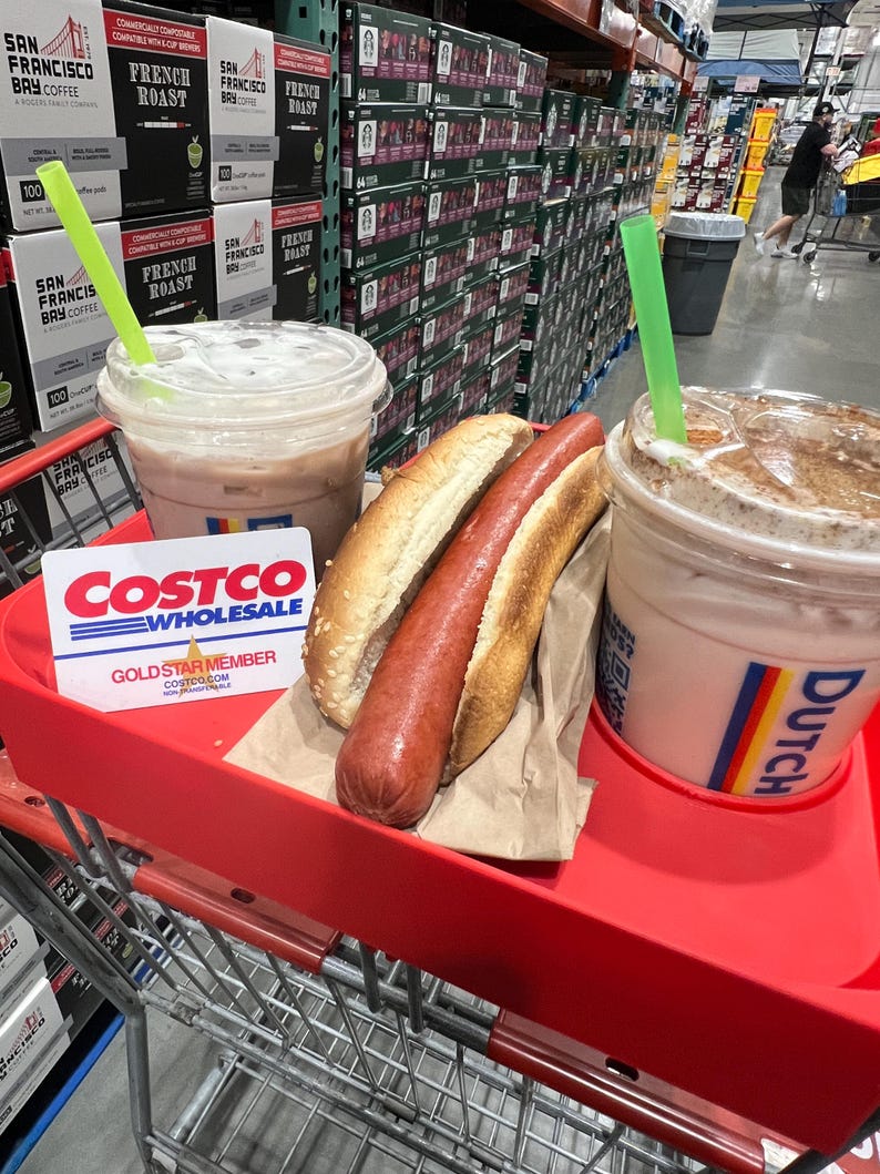 May include: A red shopping cart holds two iced coffee drinks with green straws, a hot dog in a bun, and a Costco Gold Star Member card. Coffee boxes are stacked in the background.