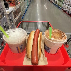 May include: A red shopping cart holds a hot dog in a bun and two iced beverages with green straws. The cart is in a large warehouse store with tall shelves stocked with merchandise.