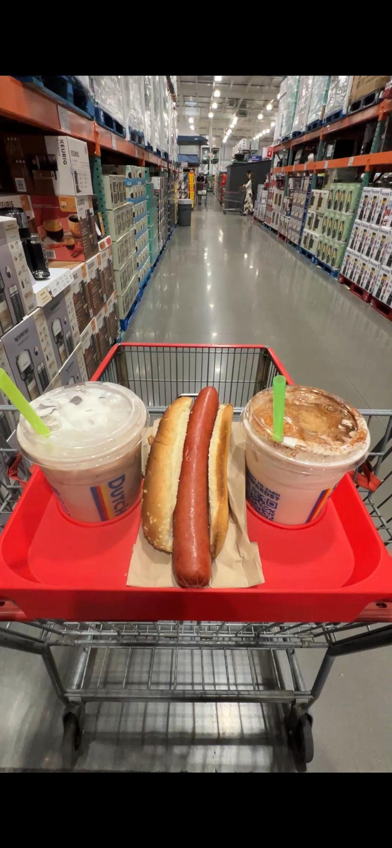 May include: A red shopping cart holds a hot dog in a bun and two iced beverages with green straws. The cart is in a large warehouse store with tall shelves stocked with merchandise.