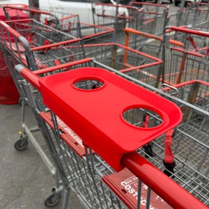 May include: A red shopping cart with a red plastic tray attached to the handle. The tray has two circular cup holders. The cart is in a parking lot with other shopping carts.