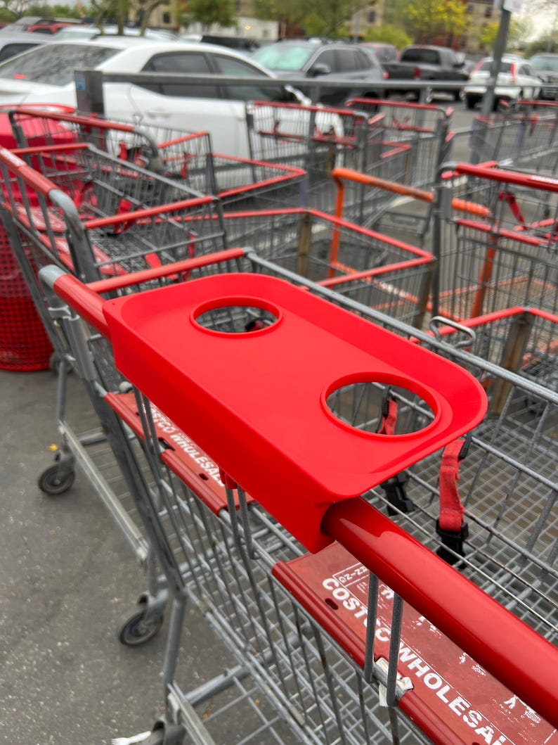 May include: A red shopping cart with a red plastic tray attached to the handle. The tray has two circular cup holders. The cart is in a parking lot with other shopping carts.