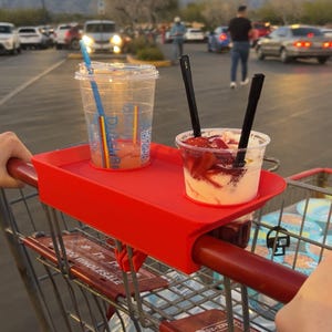 May include: A red shopping cart tray holds two clear plastic cups. One cup contains a beverage with a blue and rainbow straw. The other cup has a dessert with strawberries and two black spoons. The background shows a parking lot and mountains.