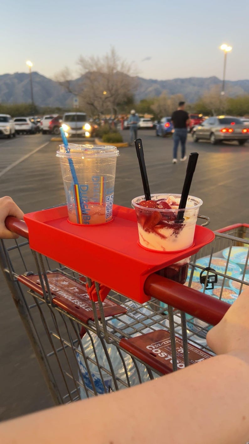 May include: A red shopping cart tray holds two clear plastic cups. One cup contains a beverage with a blue and rainbow straw. The other cup has a dessert with strawberries and two black spoons. The background shows a parking lot and mountains.