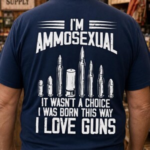 May include: A navy blue t-shirt featuring white screen-printed text that reads I'M AMMOSEXUAL, IT WASN'T A CHOICE, I WAS BORN THIS WAY, I LOVE GUNS. The design includes a graphic illustration of various bullet and shotgun shell casings in white.