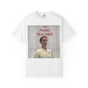 May include: White t-shirt with a movie poster design for "The Piano Teacher." The poster features a woman with pink glasses and the film's title in red. Below the image is the text "A Film by Michael Haneke."