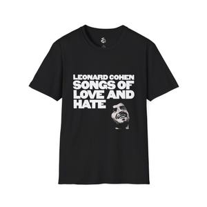 Pode incluir: Uma t-shirt preta de manga curta com o texto branco "LEONARD COHEN SONGS OF LOVE AND HATE" impresso no peito. Abaixo do texto, encontra-se um pequeno retrato fotográfico a preto e branco de Leonard Cohen a sorrir no tecido preto.