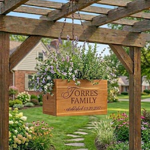 May include: A wooden planter box filled with lavender and other flowers hangs from a wooden pergola. The box is engraved with the words "Torres Family" and "established 03.20.2017". The scene is set in a well-manicured garden.