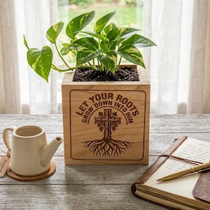 May include: A wooden planter box with a green and yellow plant. The box has the text "LET YOUR ROOTS GROW DOWN INTO HIM" with a cross and tree design. A small ceramic watering can and a journal are also visible.