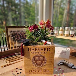 May include: A wooden planter box with the Harvard University logo, filled with a flowering plant. A miniature graduation cap and diploma are placed on top. The text "CLASS OF 2026" is visible. A framed photo and paintbrushes are in the background.