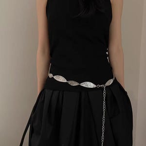 Pode incluir: Um cinto de metal prateado com um intrincado design de folhas. O cinto &eacute; usado sobre um traje preto, com uma corrente pendurada. O traje inclui um top preto e uma saia preta.