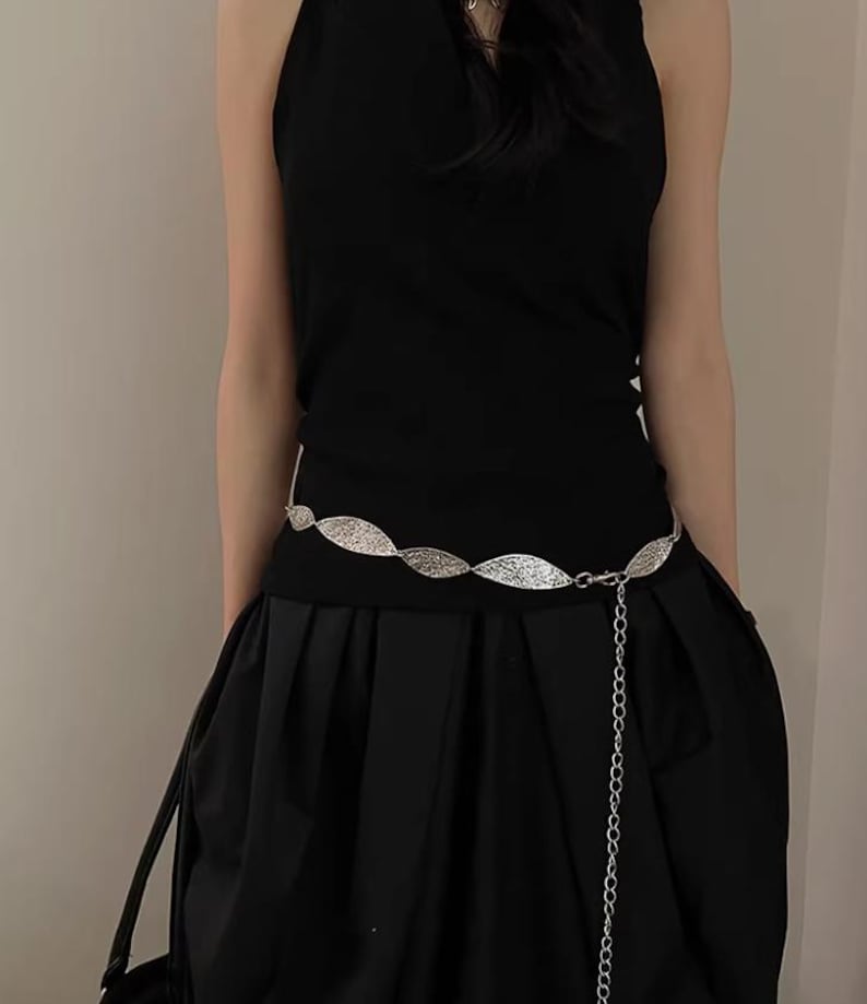Pode incluir: Um cinto de metal prateado com um intrincado design de folhas. O cinto &eacute; usado sobre um traje preto, com uma corrente pendurada. O traje inclui um top preto e uma saia preta.