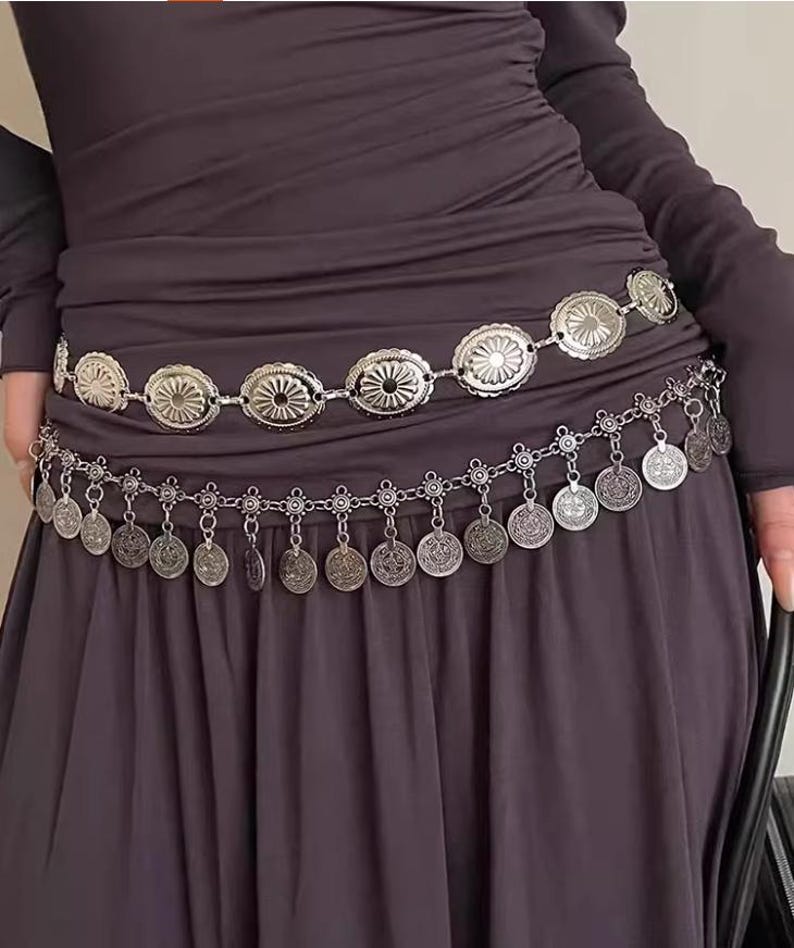 Pode incluir: Cinto de metal prateado com duas fileiras de elementos decorativos. A fila superior apresenta medalh&otilde;es ovais com um design de raios de sol, enquanto a fila inferior tem pingentes em forma de moedas. O cinto &eacute; usado sobre um vestido cinza escuro.