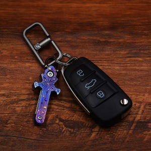May include: A keychain featuring a decorative, multi-coloured knife-shaped pendant and a black car key fob. The pendant has a blue and purple colour scheme with gold accents. The key fob has three buttons with white icons.