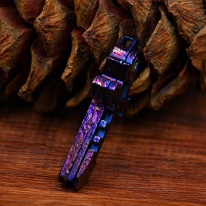 May include: A small, multi-coloured metal tool with a textured surface. The tool has a rectangular shape with a series of square cutouts and a keyhole-shaped opening. The colour shifts from blue to purple, with hints of gold.