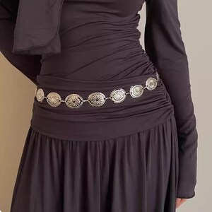 Pode incluir: Cinto de metal prateado com uma s&eacute;rie de medalh&otilde;es circulares com padr&atilde;o de raios de sol. O cinto &eacute; usado sobre um vestido escuro de manga comprida. Os medalh&otilde;es s&atilde;o ligados por pequenas elos, criando um acess&oacute;rio flex&iacute;vel e decorativo.