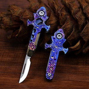 Op de afbeelding: Twee kleine, decoratieve vouwmesjes met sierlijke blauwe en paarse handvatten. De zilveren lemmeten contrasteren met de gedetailleerde handvatten, die een stervormig patroon hebben. De mesjes worden gepresenteerd op een houten oppervlak.