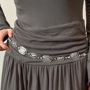 Pode incluir: Cinto de metal prateado com elos ovais e retangulares. O cinto &eacute; usado sobre um vestido cinzento, e os elos t&ecirc;m uma borda texturizada em forma de corda. Um acess&oacute;rio elegante.