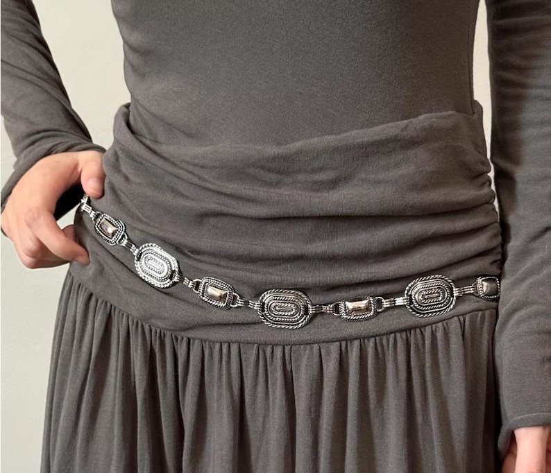 Pode incluir: Cinto de metal prateado com elos ovais e retangulares. O cinto &eacute; usado sobre um vestido cinzento, e os elos t&ecirc;m uma borda texturizada em forma de corda. Um acess&oacute;rio elegante.